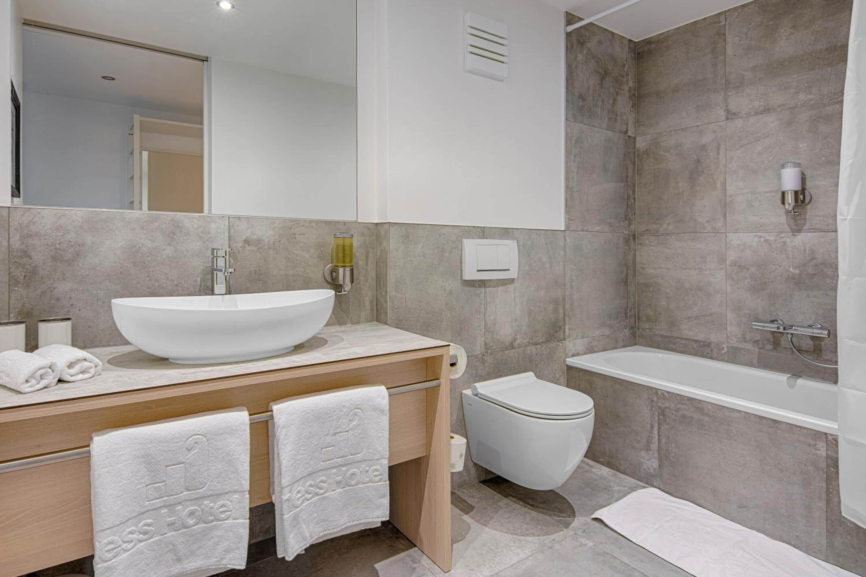 A modern bathroom with a large white sink, a toilet, and a bathtub. There are towels on the counter and a mirror above the sink.