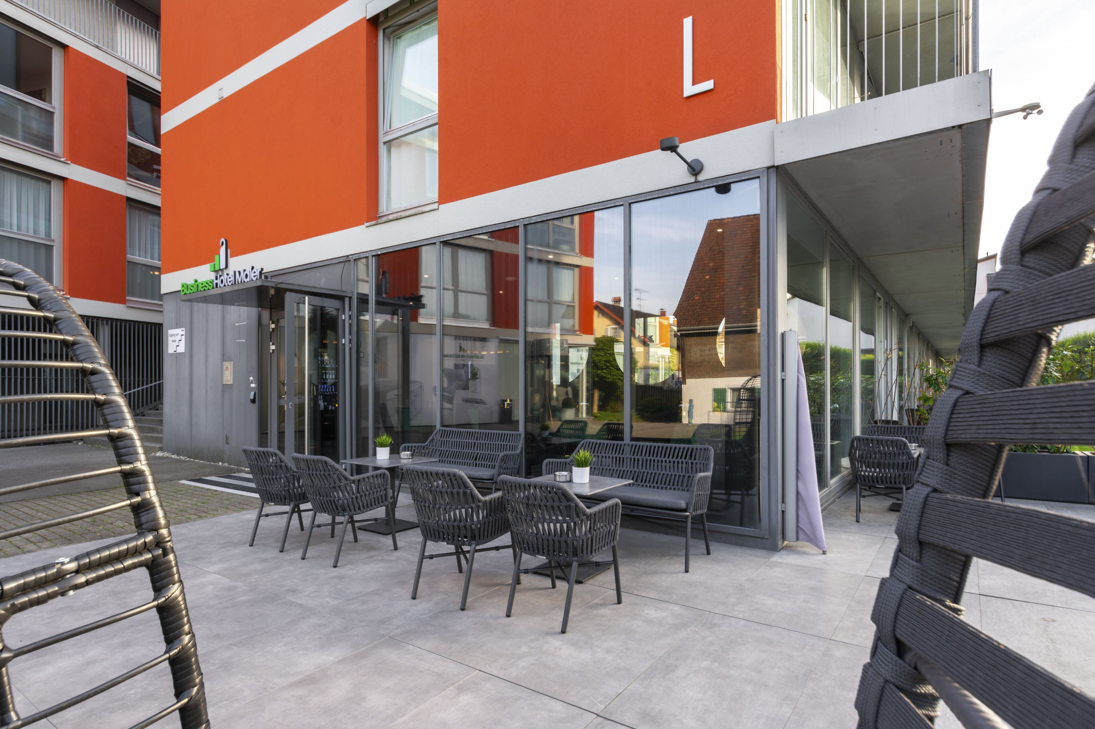 Un espace extérieur aménagé avec des tables et des chaises, une entrée avec une porte vitrée et un bâtiment dont les murs extérieurs sont peints en orange et en blanc.