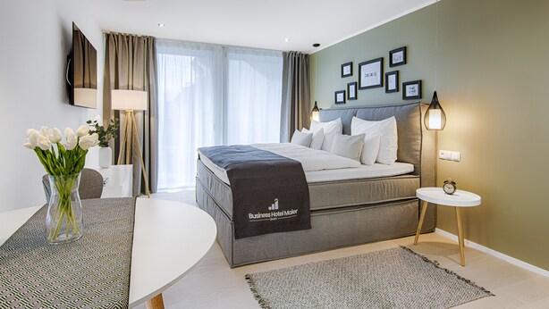 A modern hotel room with a large bed, a bedside table, a lamp, and a small table with a clock.