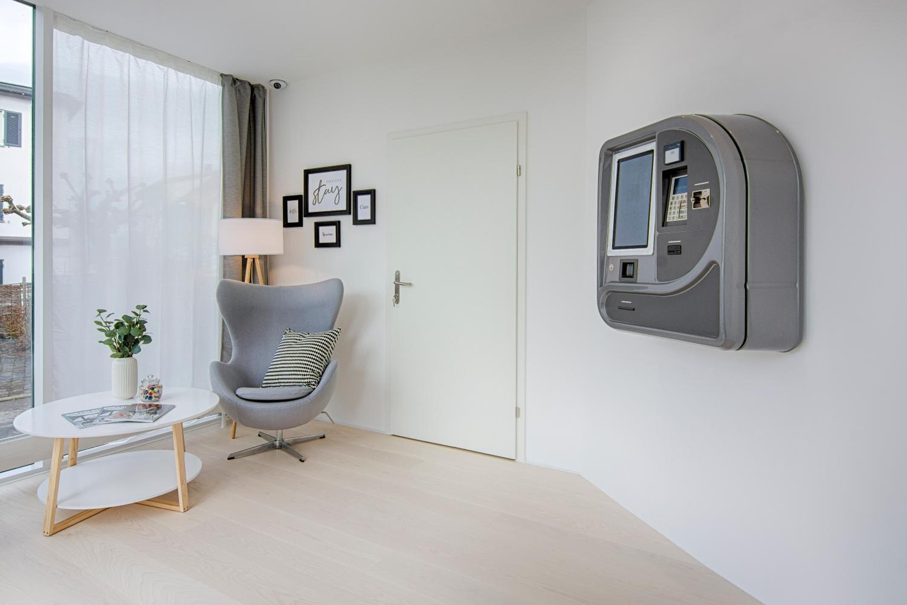 Una habitación moderna con un sillón gris, una mesita blanca pequeña, una lámpara y un televisor montado.