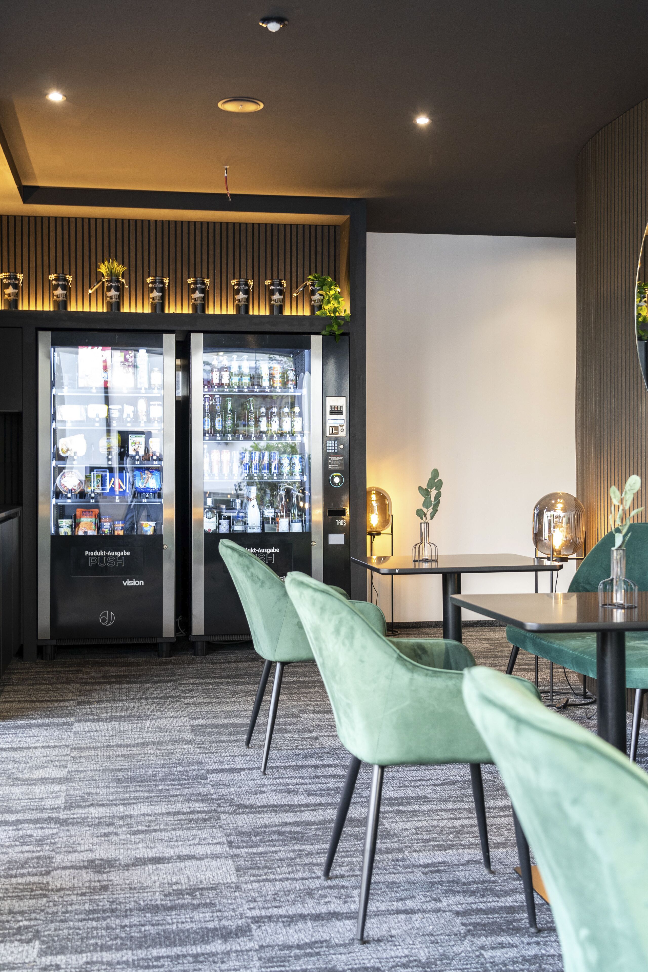 Een moderne bar met twee automaten, gelabeld 'Product-Ausgabe PUSH vision', een tafel met stoelen en een groot scherm.