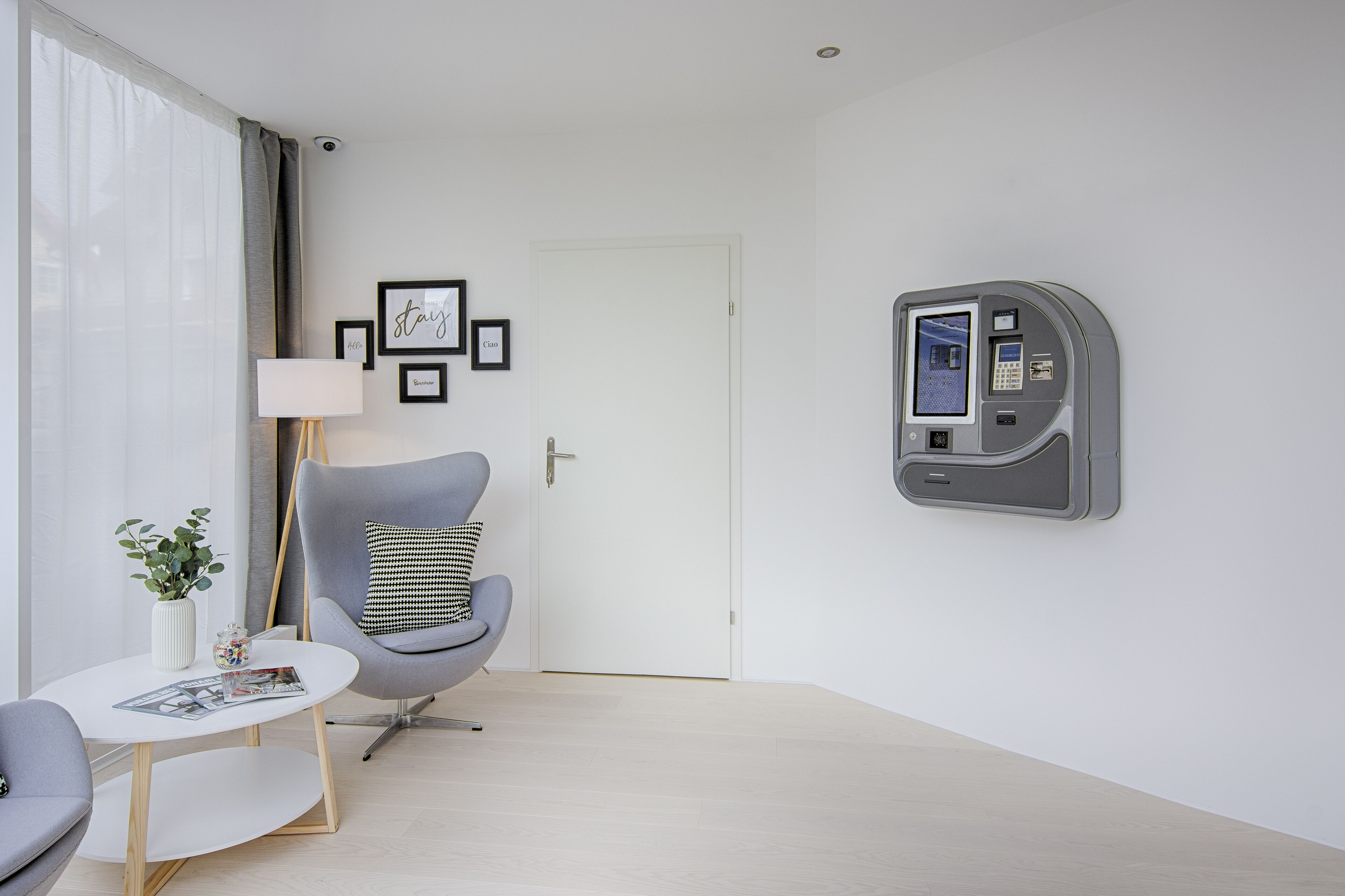 Habitación de hotel moderna con un sillón gris, una mesita blanca con una revista, una planta en maceta y un panel de control en la pared.