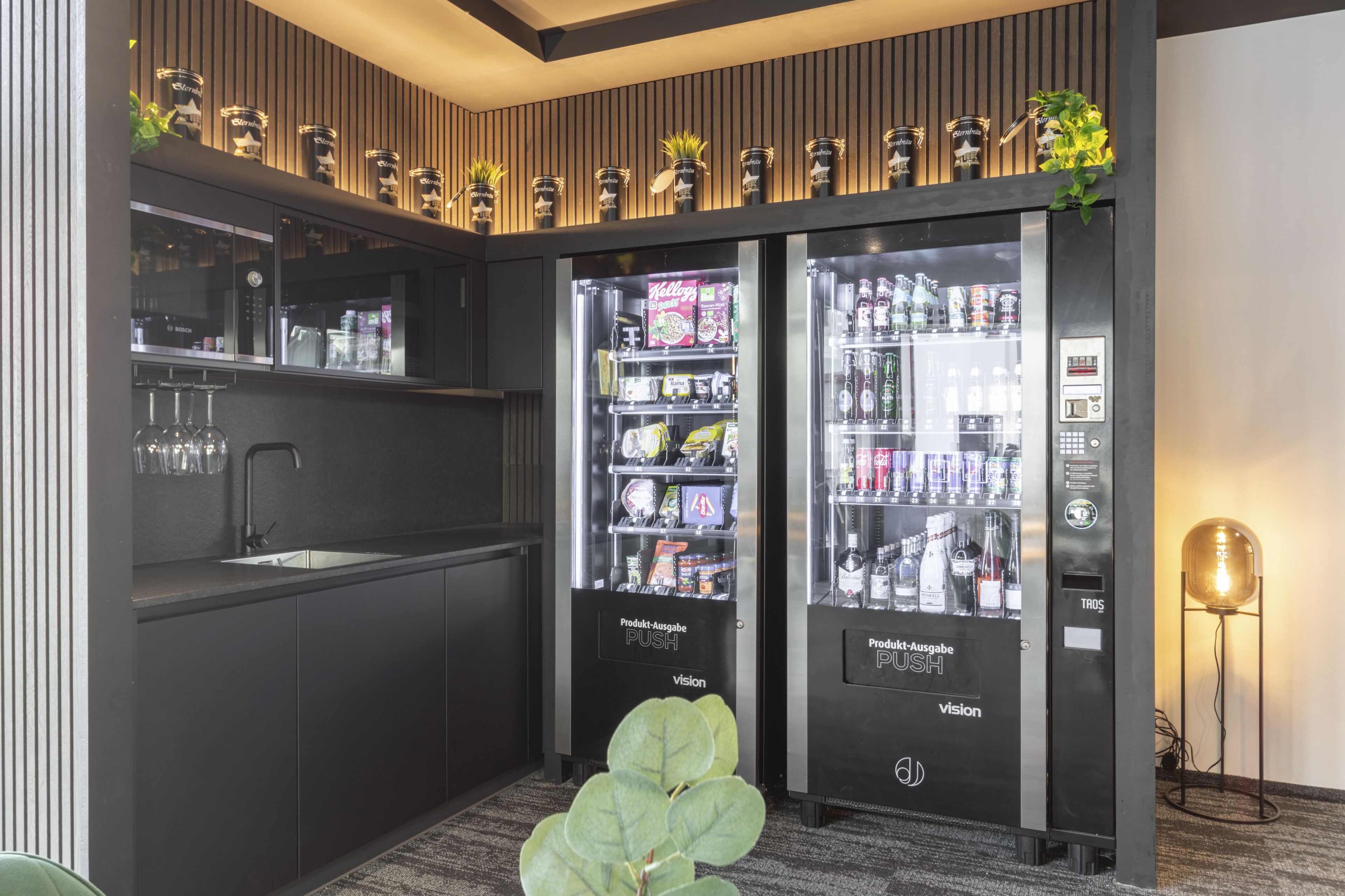 A modern kitchen with a sink, two beverage vending machines, and a potted plant.