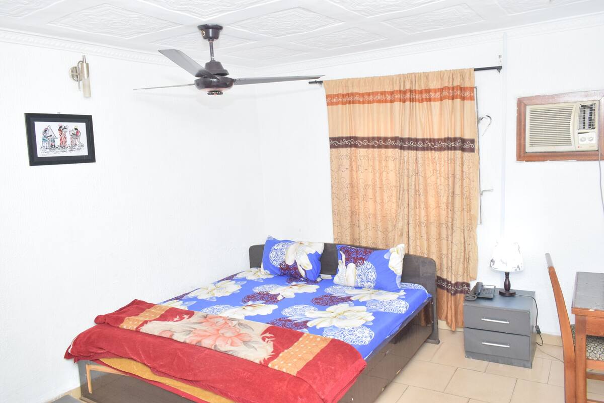A bedroom with a bed, a ceiling fan, a nightstand, a desk, and a window with curtains.