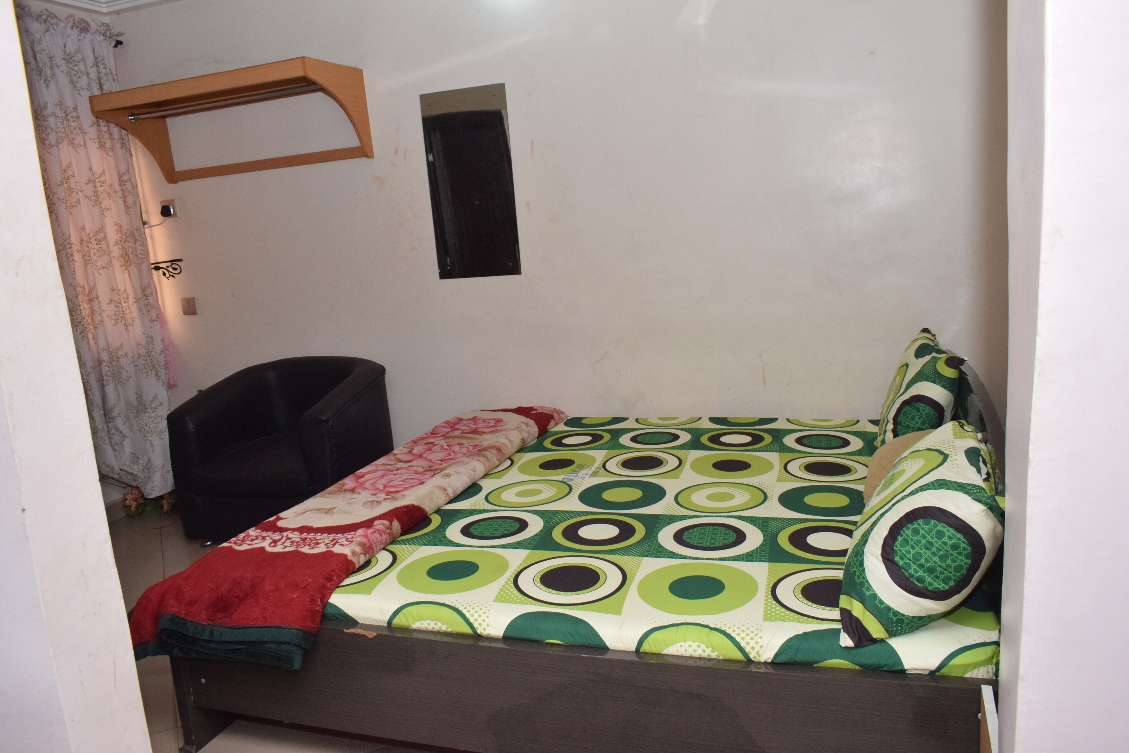 Une petite chambre avec un lit simple, une tête de lit en bois, une couvre-lit à motifs, un plaid rouge et une chaise noire.