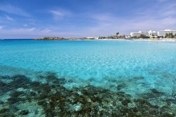 A beach with clear turquoise water, a rocky seabed, and a distant shoreline with buildings.