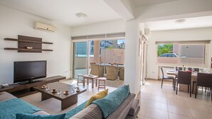 A modern living room with a flat-screen TV, a sofa, a coffee table, and a dining area with chairs and a table.
