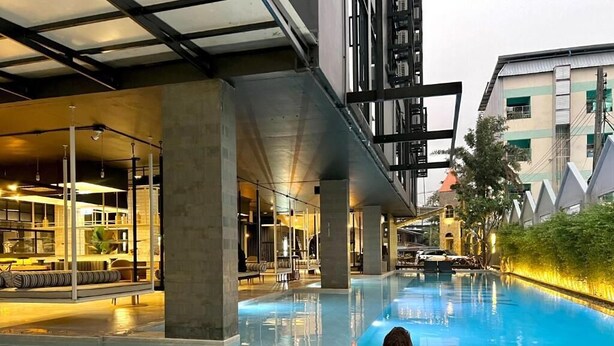 A person relaxing in a lounge chair by a pool with a bar area and indoor seating visible in the background. - BOOK Design Hotel (Chiang Mai)