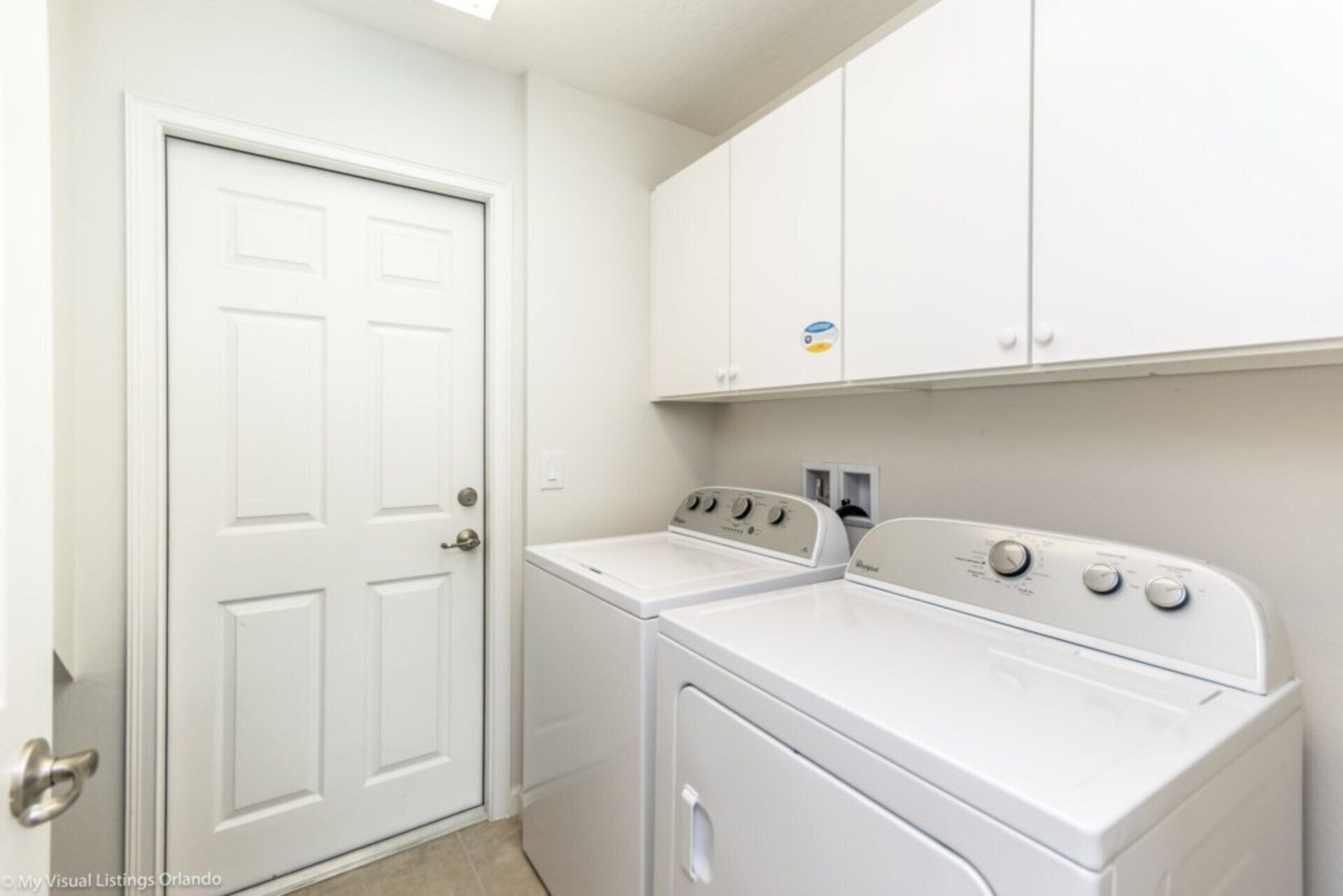 A laundry room with a white door, two stacked washers and dryers, and white cabinets above.