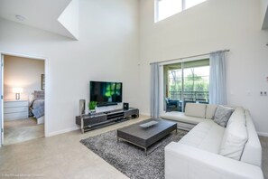 Ein modernes Wohnzimmer mit Flachbildfernseher, einer Couch, einem Couchtisch und einem großen Fenster mit Vorhängen.