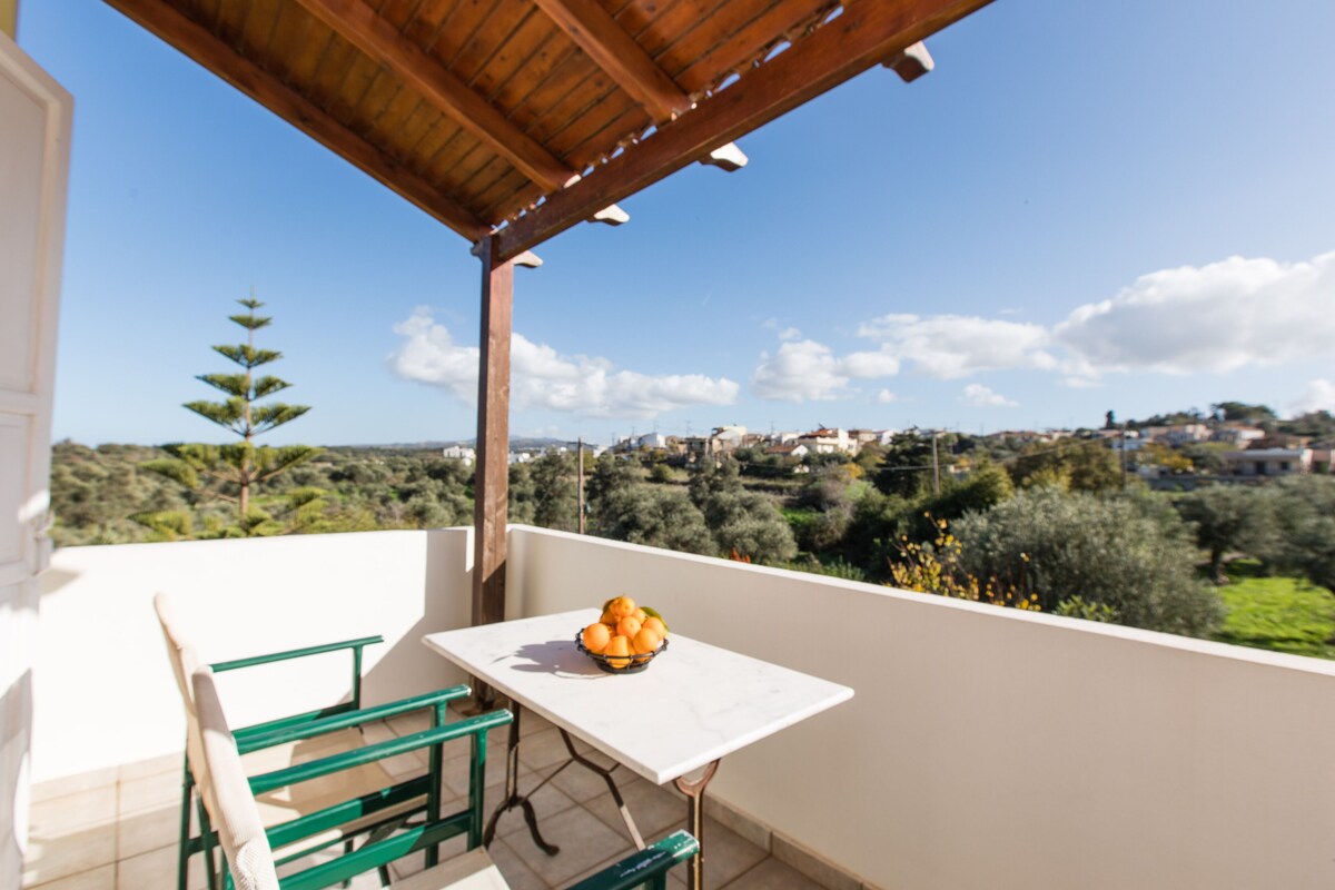 Un balcon avec une table et des chaises, un saladier rempli de fruits et une vue sur un paysage verdoyant, avec des arbres et des bâtiments.