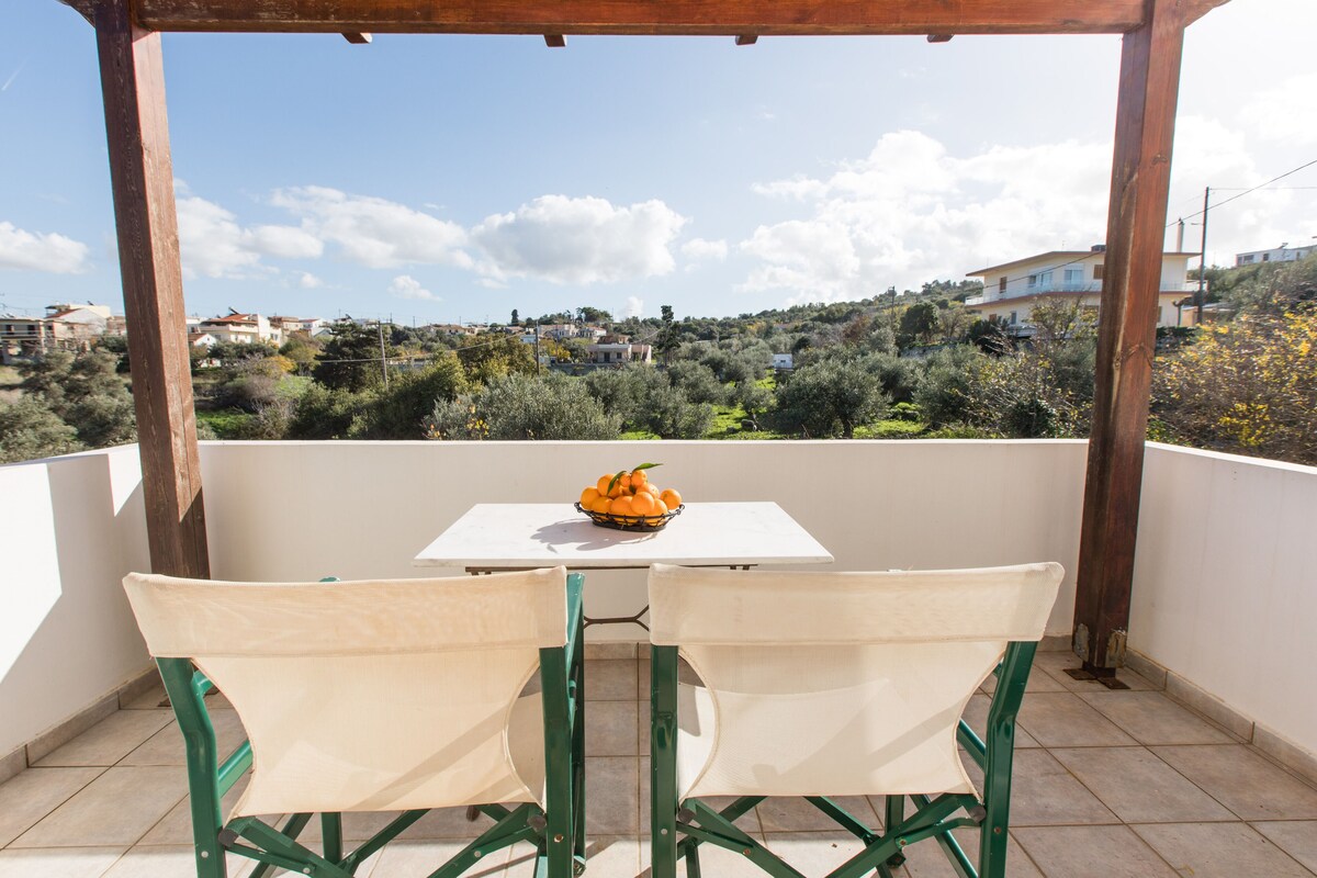 Un balcon avec deux chaises et une table, un saladier rempli d’oranges et une vue sur un paysage vallonné parsemé de bâtiments.