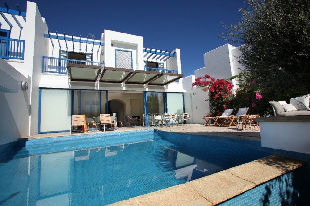 A modern pool area with clear blue water, white walls, and blue accents. There are lounge chairs and a covered seating area.