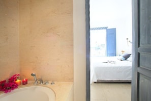 A bathroom with a bathtub, a sink, and a mirror. There is a bed visible through an open door in the adjacent room.