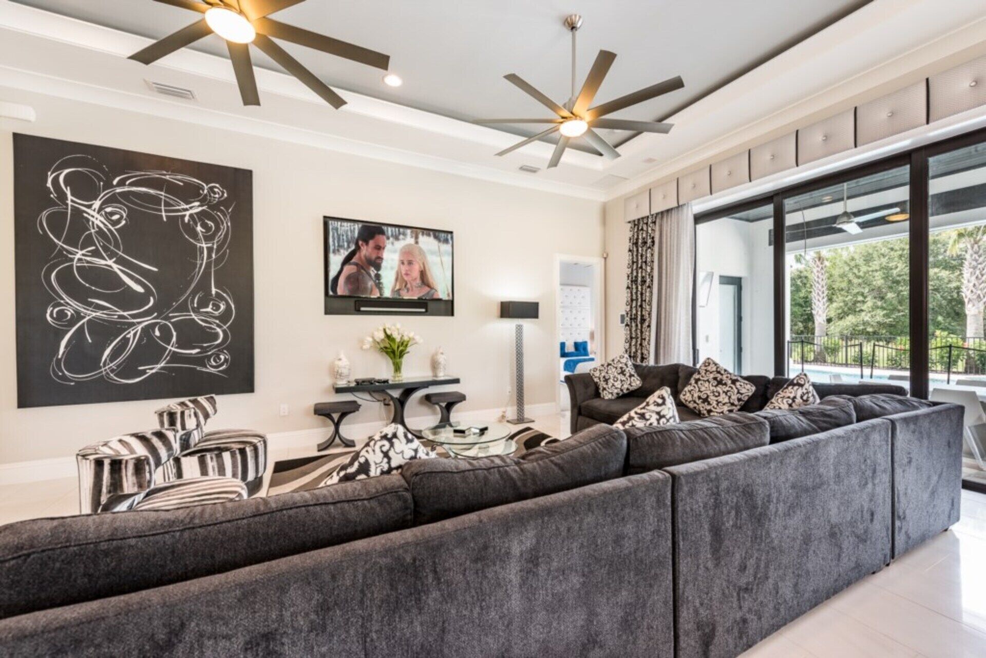 Ein modernes Wohnzimmer mit einem großen Ecksofa, Deckenventilatoren, einem Flachbildfernseher und einem großen Fenster mit Blick ins Freie.