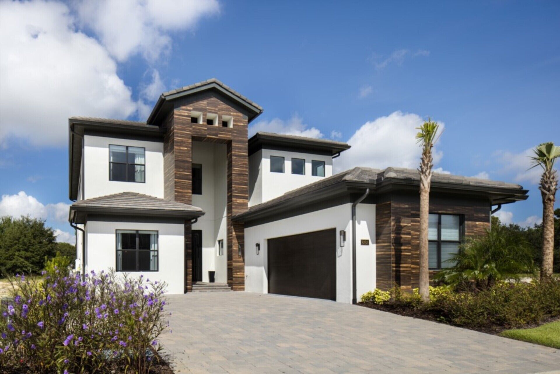 A modern two-story house with a garage, a covered driveway, and a landscaped front yard with purple flowers.