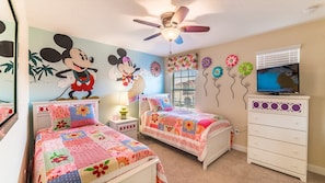 Kinderzimmer mit zwei Betten, einem Deckenventilator, einem Fernseher und Wanddekorationen, darunter Mickey Mouse und Blumen.