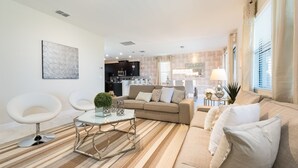 Un salon moderne avec un canapé, une table basse et des fauteuils. On aperçoit une cuisine au fond.