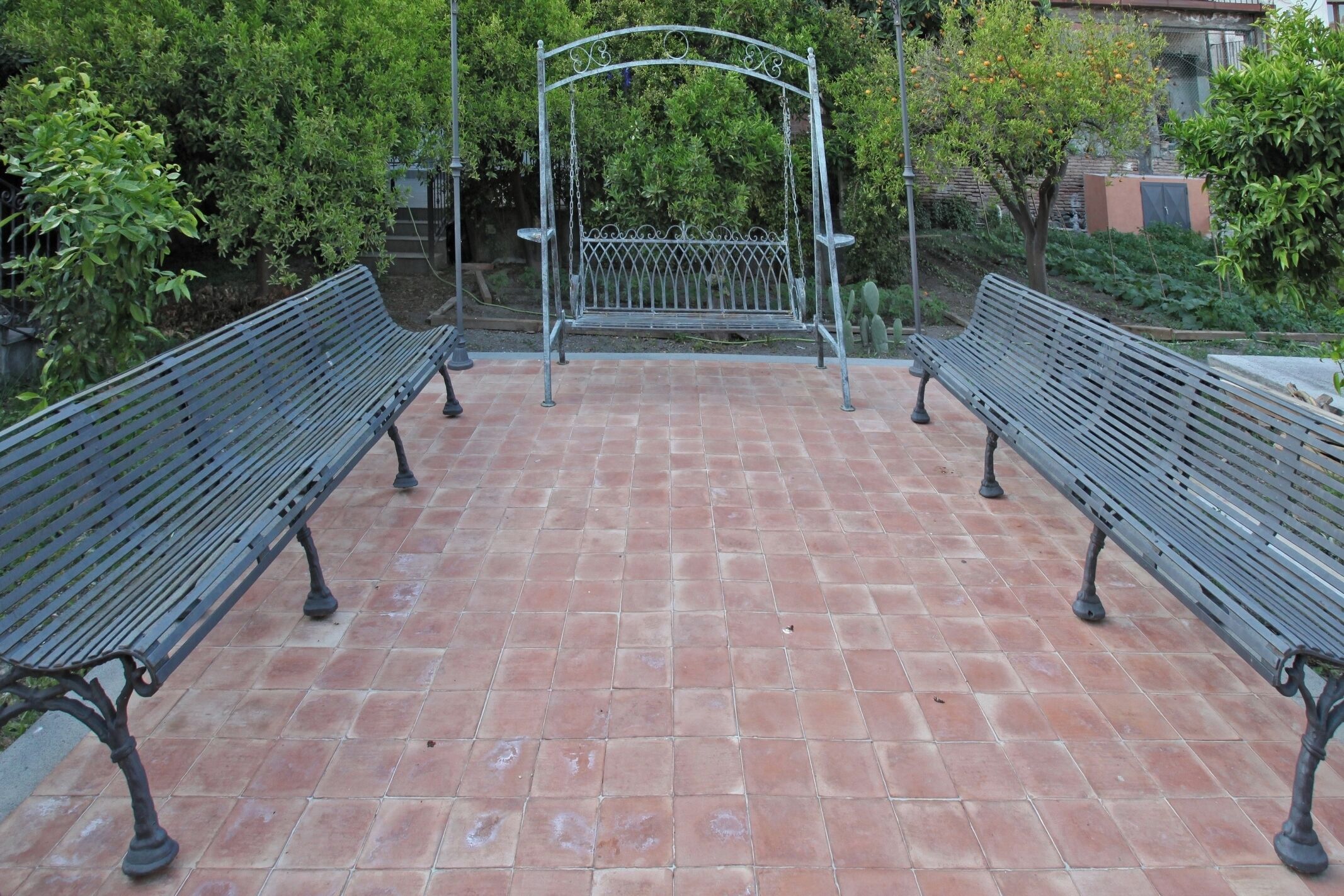 A paved patio with two metal benches and a wrought iron swing set.