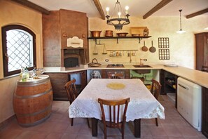 Uma cozinha rústica com um barril de madeira, uma mesa com toalha rendada e um lustre.
