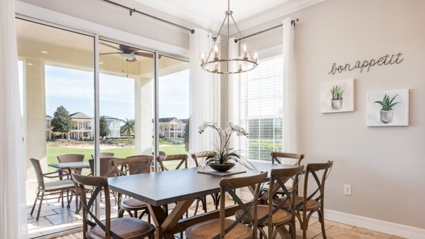 Un espace repas comprenant une grande table et des chaises, un lustre, et donnant sur un quartier résidentiel.
