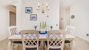 Un coin repas avec une table et des chaises blanches, un lustre, des tableaux encadrés et une horloge au mur.