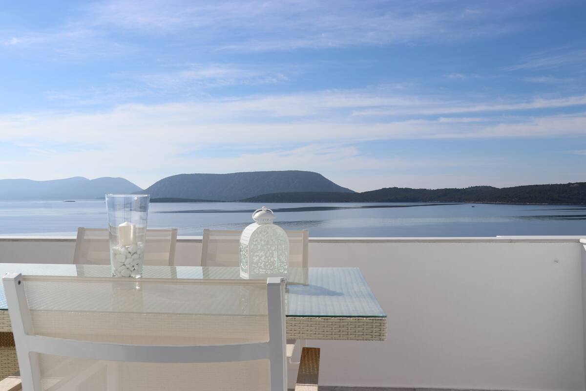 A table with a glass vase and a bottle, overlooking a calm body of water and distant mountains.