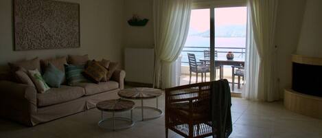 A living room with a sofa, two round tables, a wicker chair, a fireplace, and a view of the sea through a large window.