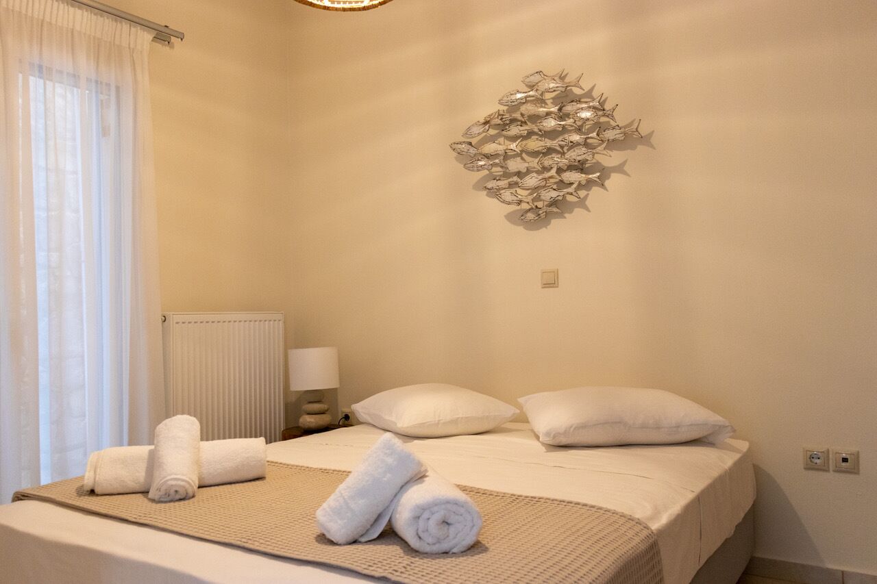 Hotellihuone, jossa on sänky, kaksi tyynyä, seinätaideteos, ikkuna verhoilla ja yöpöytälamppu.