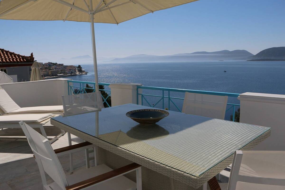 A balcony with a table and chairs, overlooking a body of water and distant mountains.