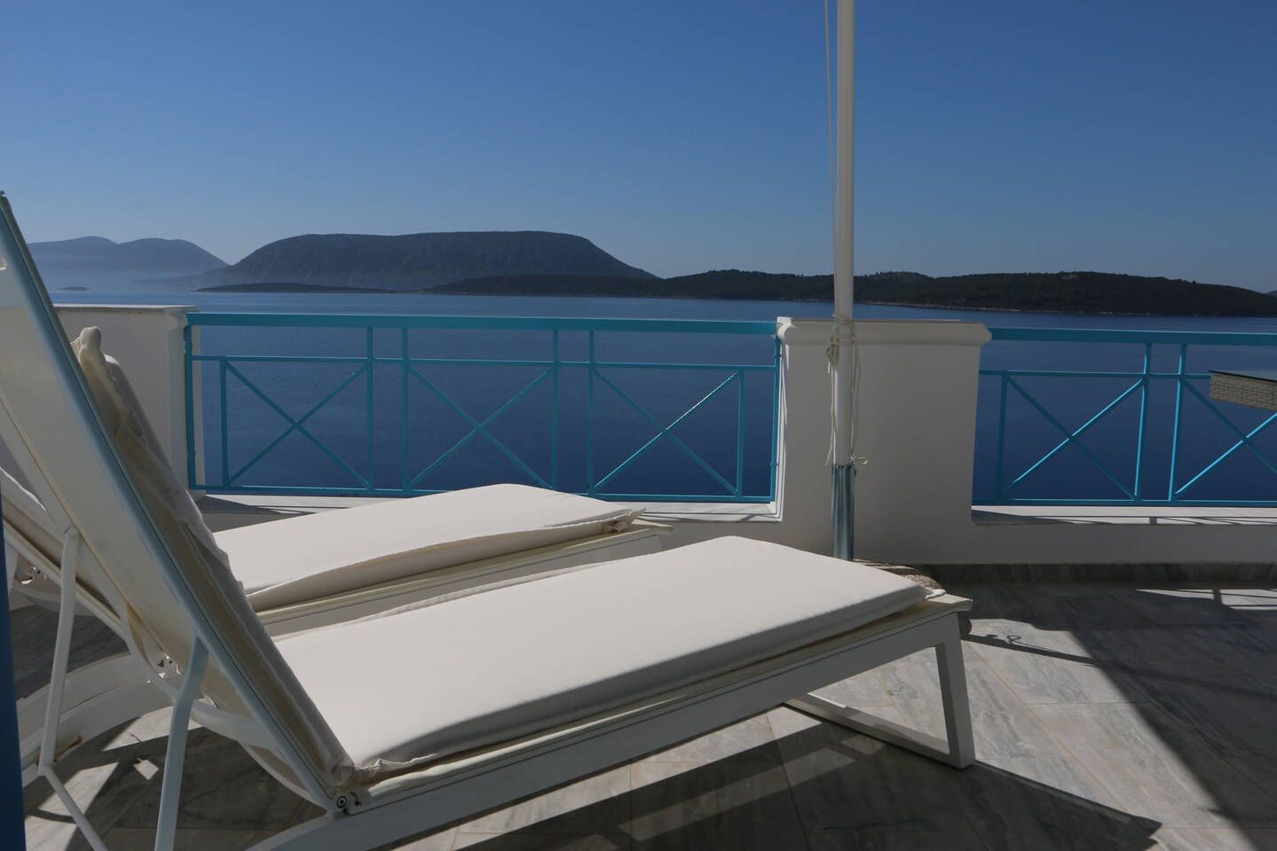 A sunlit balcony with two white lounge chairs, a blue railing, and a view of the sea and distant hills.
