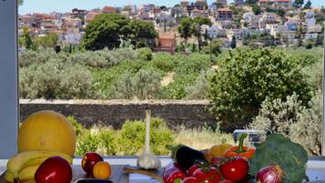 A table with fruits and vegetables in front of a window overlooking a hillside with buildings.