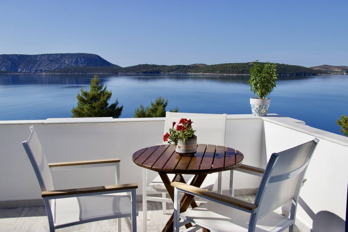 A round wooden table with two white chairs, a potted plant, and a vase with red flowers on a balcony overlooking a calm lake with distant hills.