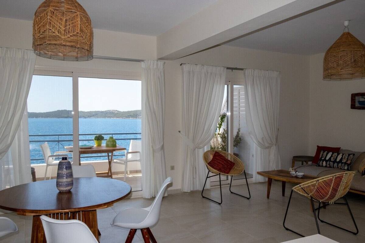 A spacious living area with a view of the sea, a round wooden table, white chairs, a sofa with cushions, and wicker chairs.