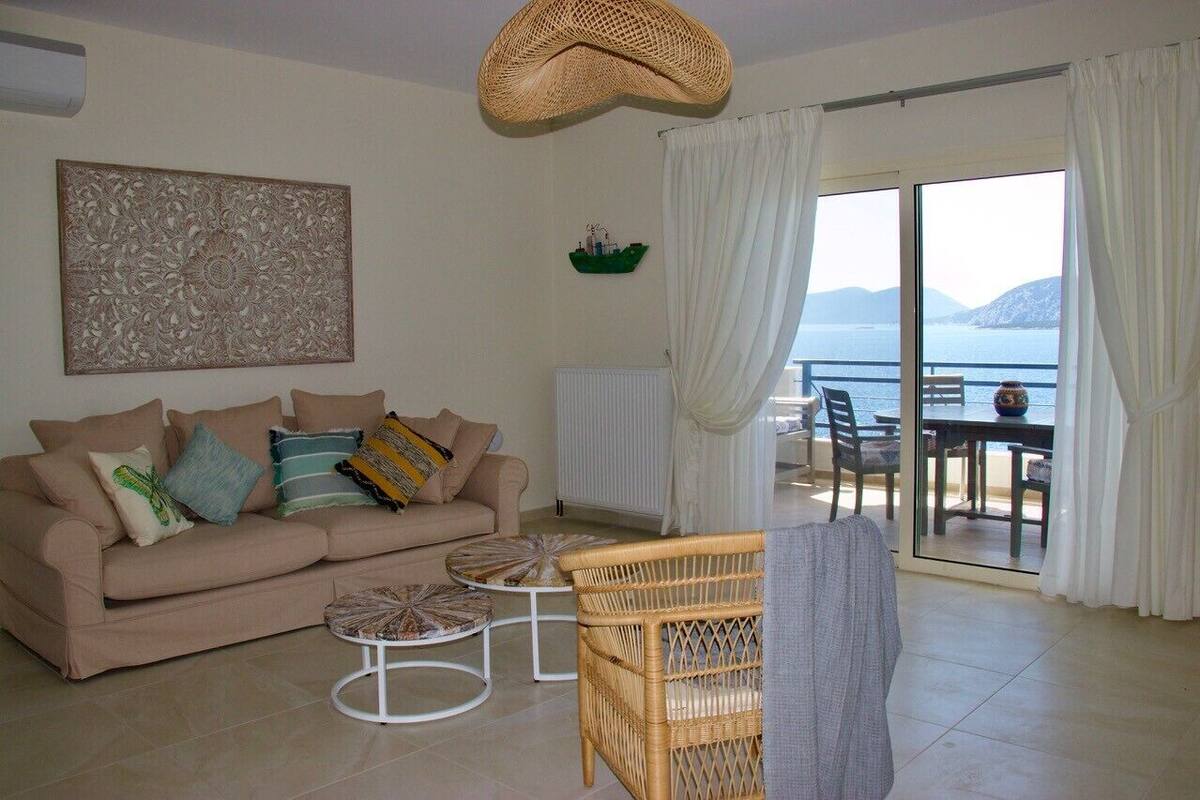 A living room with a sofa, two round tables, a wicker chair, a decorative wall piece, and a view of the sea through a large sliding glass door.
