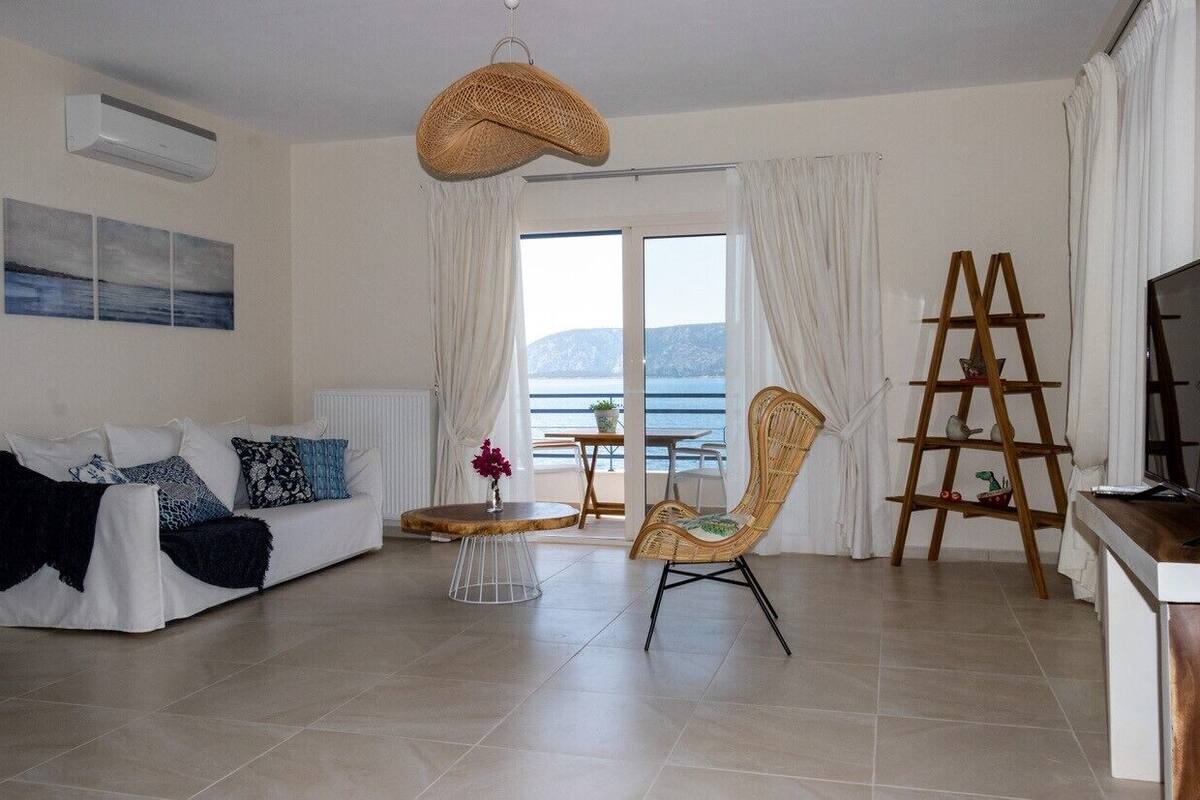 A living room with a sofa, a chair, a table, and a TV stand. There is a window with curtains and a view of the sea.