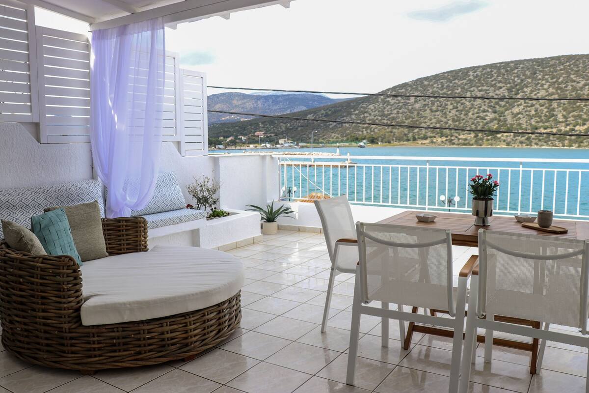 A balcony with a wicker sofa, white chairs, and a table with a view of the sea and mountains.
