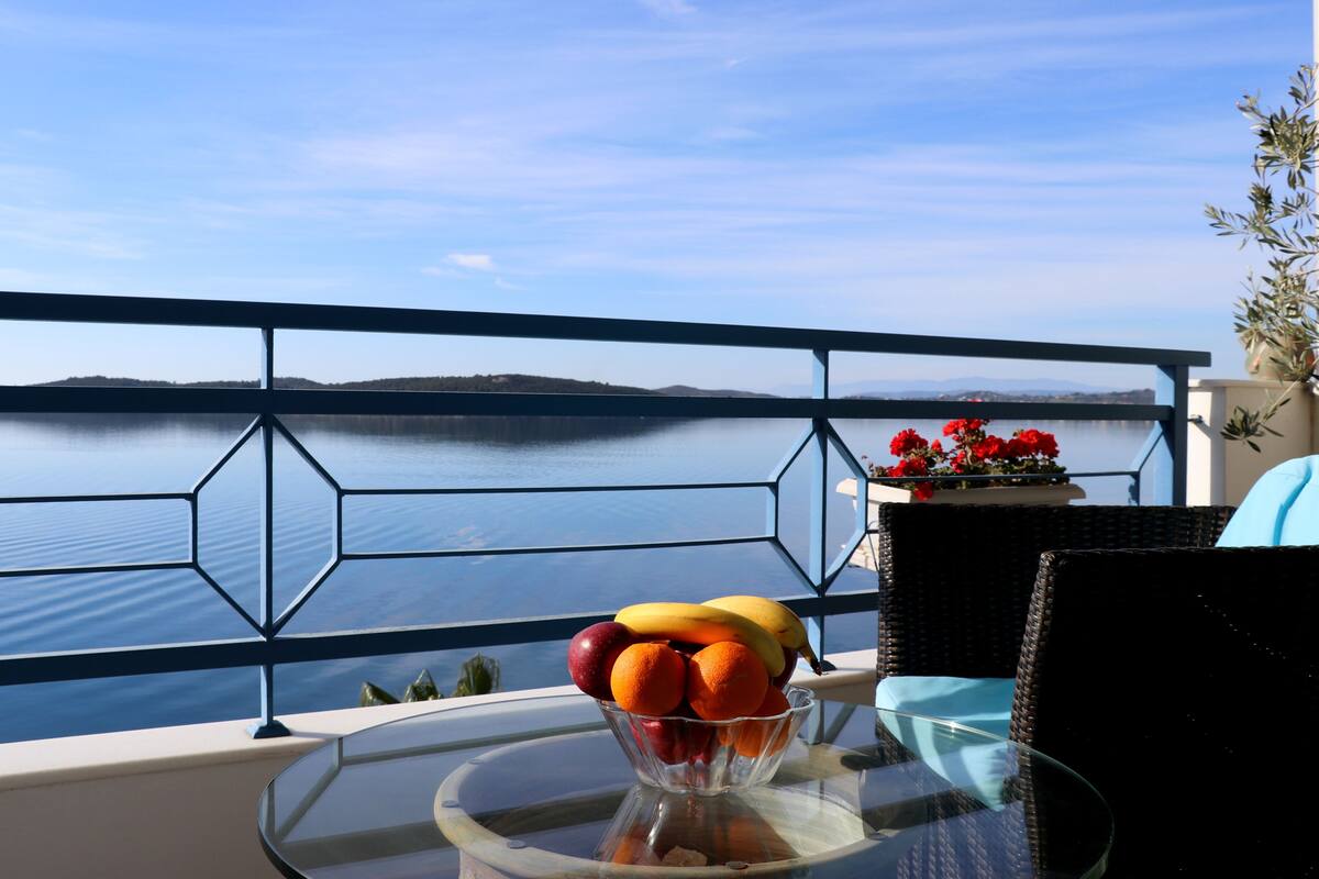 A balcony with a glass table holding a fruit bowl, a wicker chair, and a potted plant with red flowers.