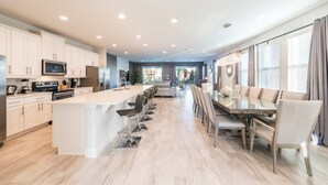 A modern kitchen with a central island, a dining area with a long table, and a view of the outdoors through large windows.
