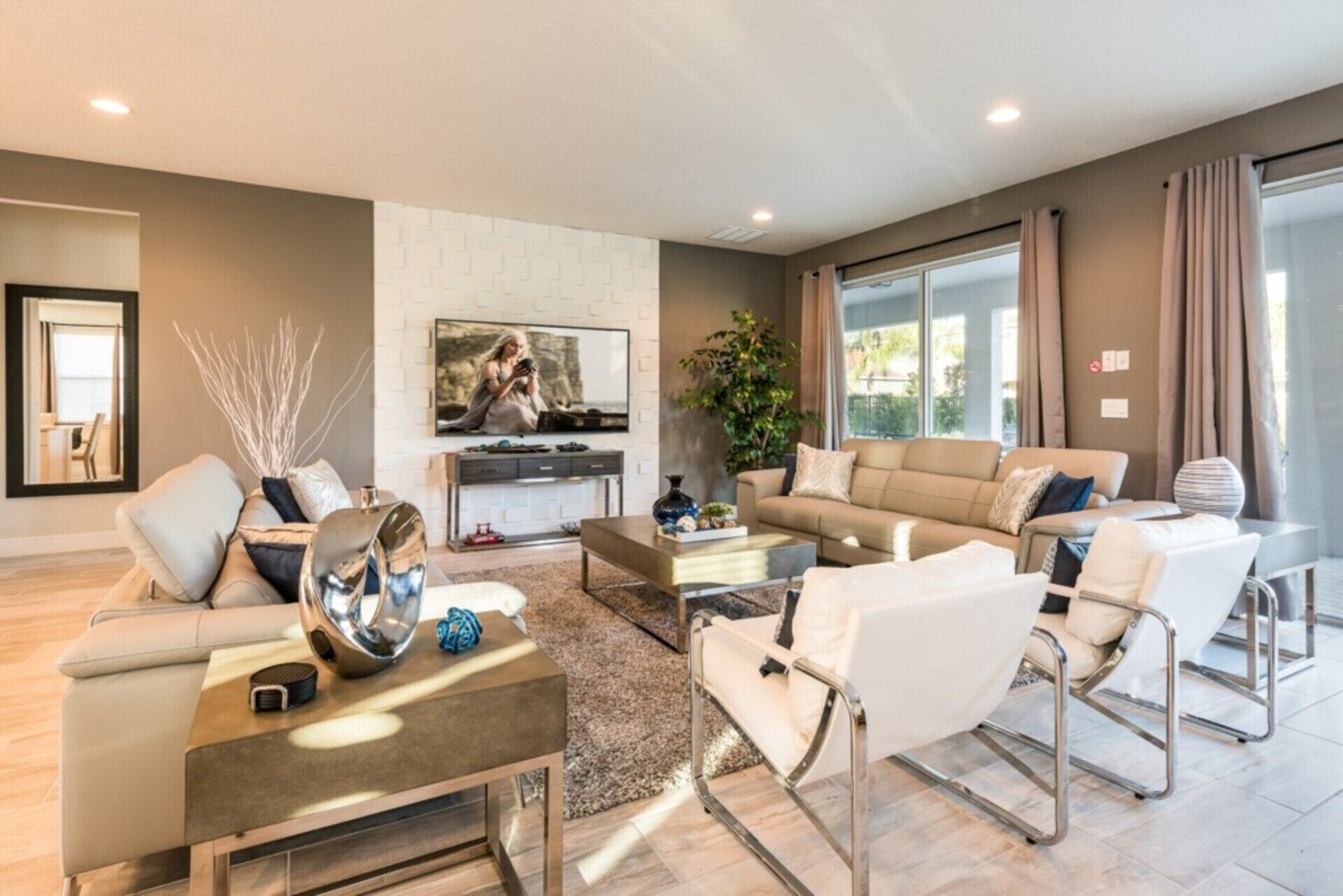 A modern living room with a sofa, coffee table, and chairs. There is a TV mounted on the wall and a plant in the corner.