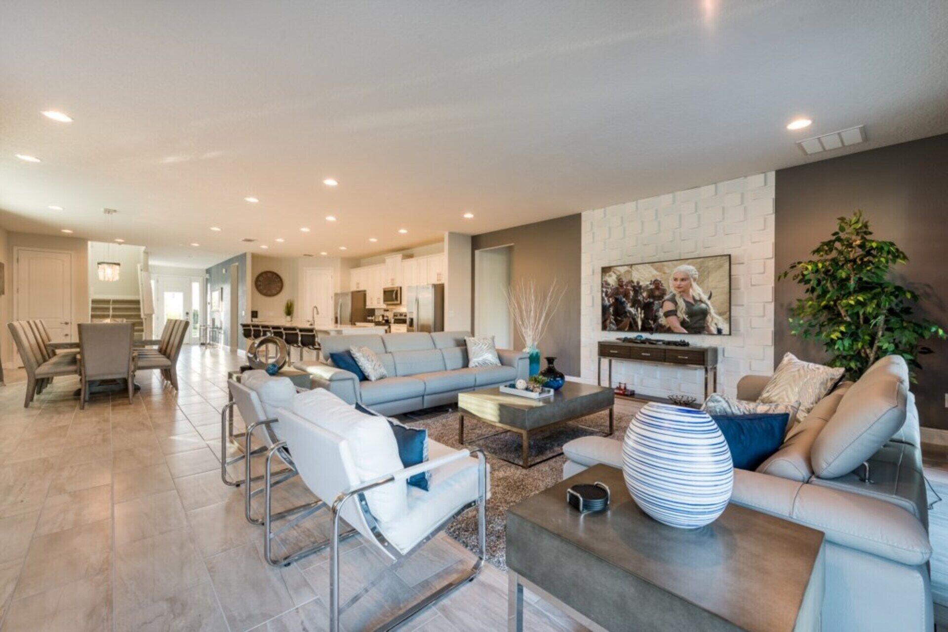 A spacious living room with a grey sofa, white armchairs, and a coffee table. There is a kitchen in the background with white cabinets and a dining area with a table and chairs.