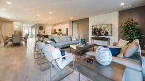 A spacious living room with a grey sofa, white armchairs, and a coffee table. There is a kitchen in the background with white cabinets and a dining area with a table and chairs.