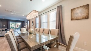 A modern dining area with a glass table, cushioned chairs, a chandelier, and a large window with blinds.