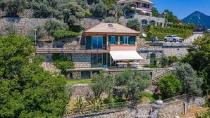 Uma casa de pedra de dois andares com varanda, rodeada por vegetação e com montanhas ao fundo.