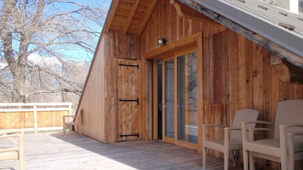 Una cabaña de madera con techo inclinado, una terraza con mobiliario exterior y un ventanal corredero que da al interior.