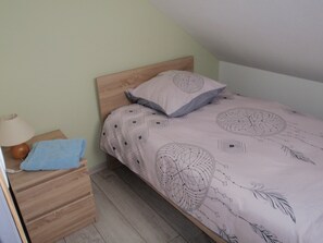 Una cama individual con una colcha estampada, cabecera de madera, lámpara de noche y una toalla azul sobre una mesita de noche de madera.