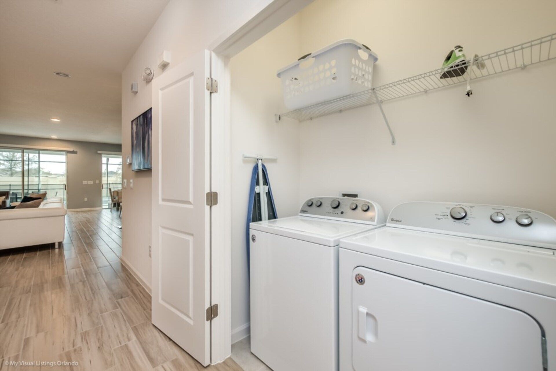 A laundry room with a washer and dryer, a shelf with laundry baskets, and a door leading to a living area.
