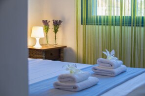 Ein Hotelzimmer mit Bett, Nachttisch mit Lampe, zwei Vasen mit Blumen und einem Fenster mit vertikalen Jalousien.