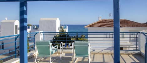 A terrace with two green chairs and a table, overlooking a coastal view with buildings and the sea.