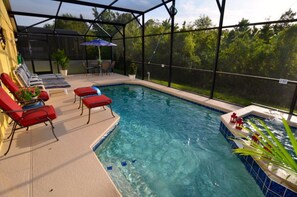 An indoor swimming pool with red lounge chairs, a covered patio area, and a view of trees through a glass enclosure.